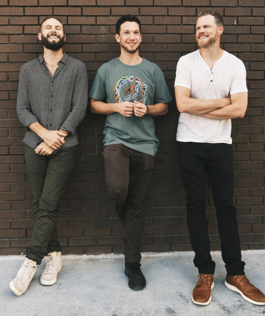 Lemon City Trio members smiling against a brick wall - Nick Tannura with guitar, Brian Robertson with keyboards, and Aaron Glueckauf with drums.