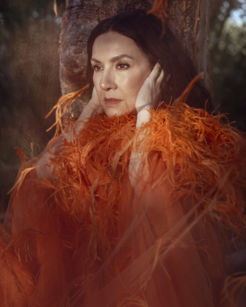 Roxana Amed, an award-winning Argentine-American jazz vocalist and composer, posed in a vibrant orange feathered outfit against a tree. Her intense gaze and contemplative expression highlight her artistic depth and the rich textures of her ensemble. Known for her unique voice and innovative jazz compositions, Amed has received critical acclaim and prestigious awards throughout her career. This image captures her as a captivating figure in the jazz music scene. Photo by Omar Cruz.
